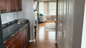 Un pasillo con suelo de madera, una zona de cocina con encimeras oscuras y un salón con sofá y cortinas.