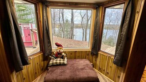 Une cabane avec un lit, une fenêtre donnant sur des arbres et un lac, et un jouet en peluche posé sur le rebord de la fenêtre.