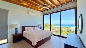 A bedroom with a large bed, two bedside tables with lamps, a ceiling fan, and a view of the ocean.