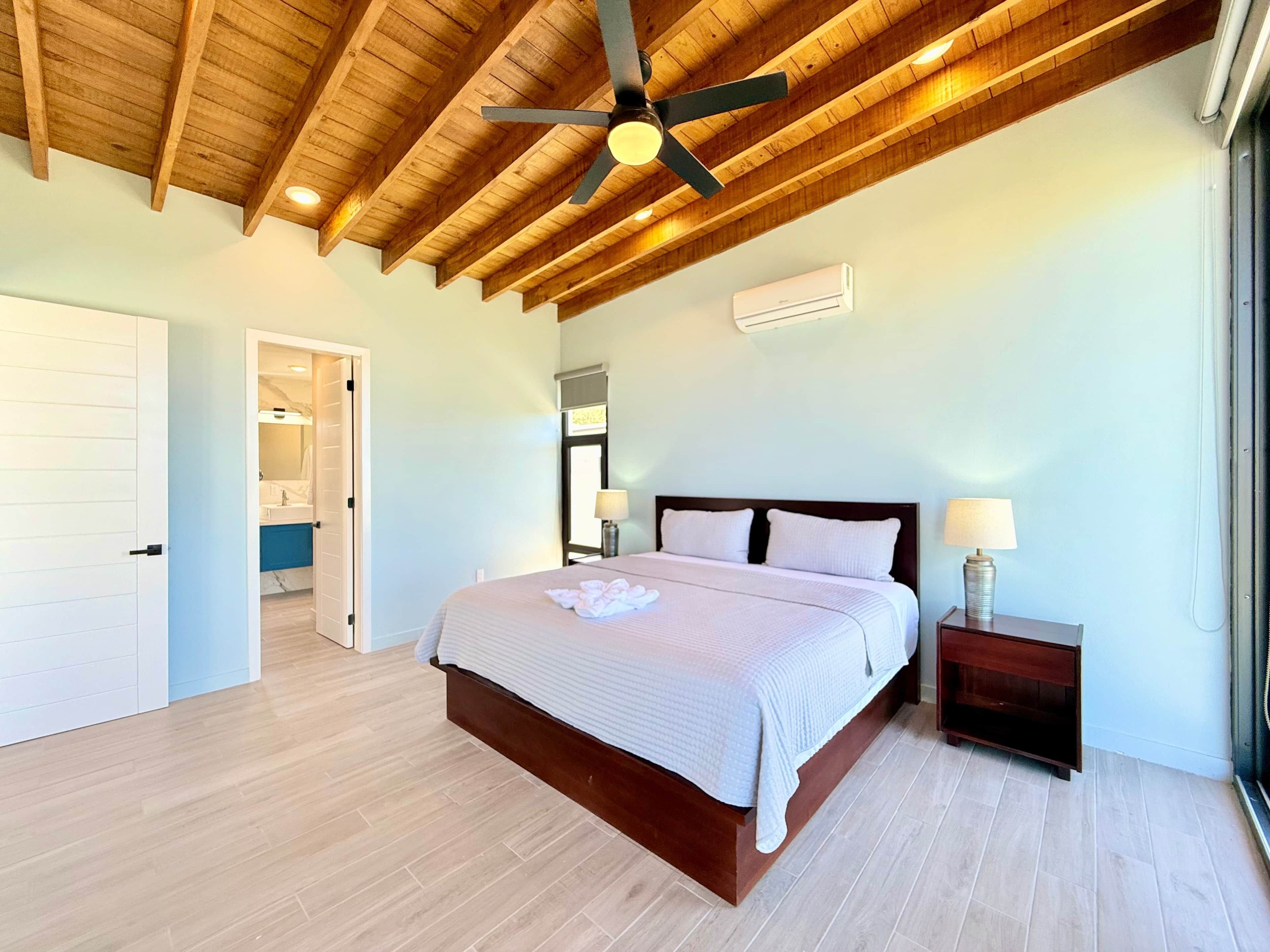 A bedroom with a wooden ceiling, a bed with white bedding, a nightstand with a lamp, and a bathroom visible through an open door.