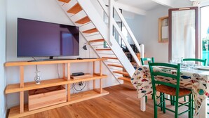 Una sala de estar moderna con un mueble bajo para televisor de madera, un televisor de pantalla plana, una escalera con escalones de madera, y un comedor con sillas verdes y una mesa cubierta con un mantel estampado con motivos de peces.