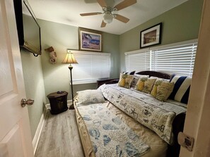 Une chambre à coucher comprenant un lit, un ventilateur de plafond, une lampe et deux fenêtres équipées de stores.