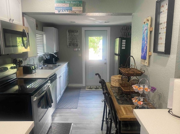 Cocina compacta con microondas, estufa y lavamanos. Hay una zona de comedor con mesa y sillas. Se ve un refrigerador al fondo.