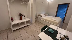 Una habitación pequeña y bien iluminada con una cama, una mesita de noche, una silla y una mesa con un florero y un vaso.