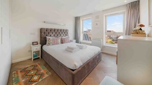 A modern bedroom with a large bed, a bedside table, a small nightstand, a window with curtains, and a view of the city.