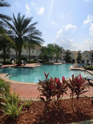 A swimming pool with a curved edge, surrounded by palm trees and lounge chairs, with buildings in the background.