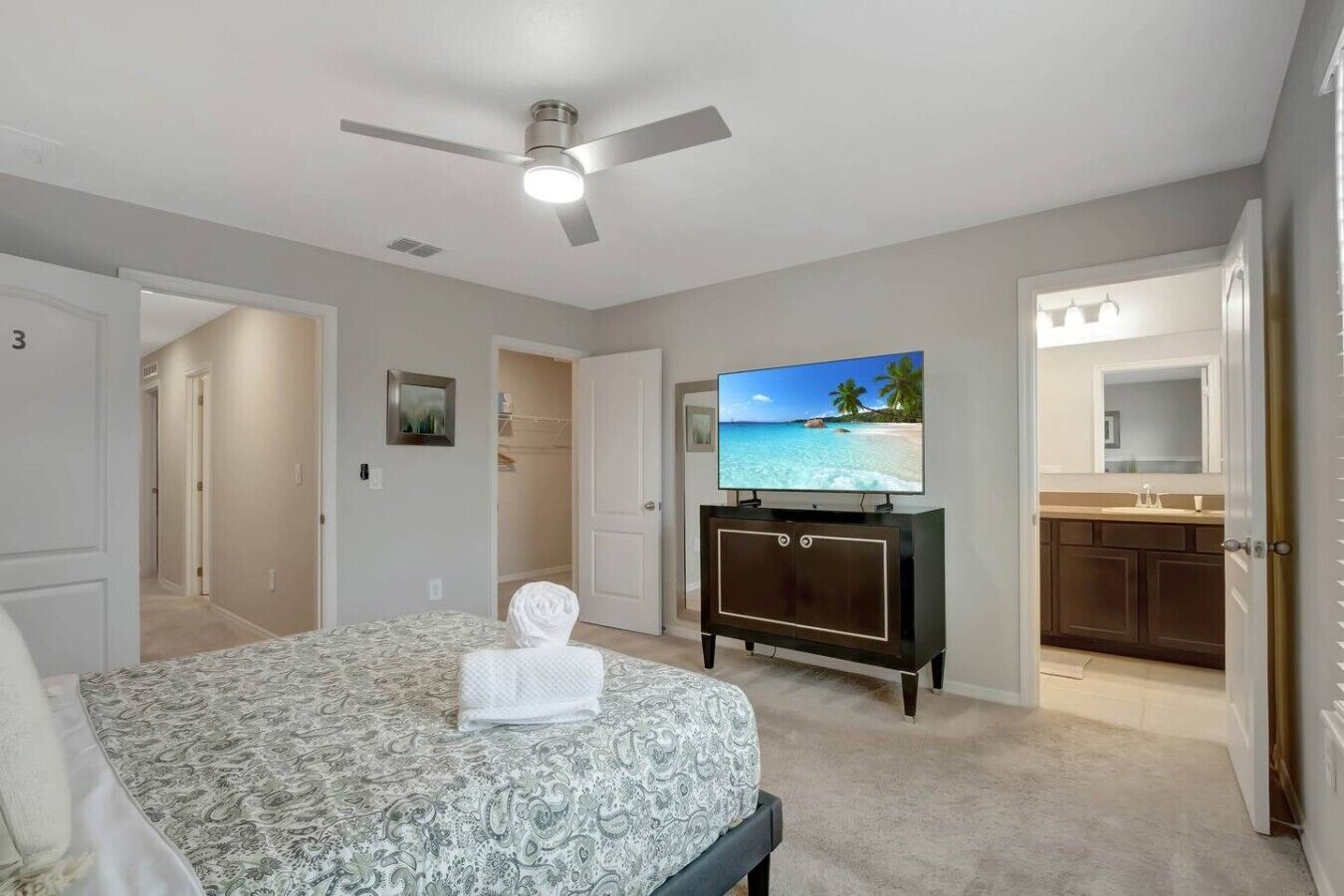 A hotel room with a bed, a television on a cabinet, a ceiling fan, and a view of a beach through the window.