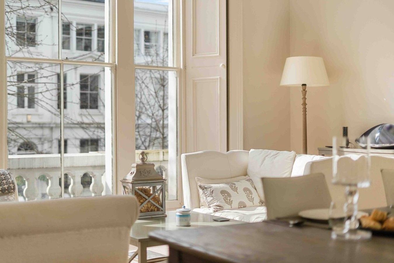 A room with a large window, a white sofa, a table with a lamp, and a view of buildings outside.