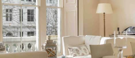 A room with a large window, a white sofa, a table with a lamp, and a view of buildings outside.
