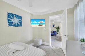 Ein Schlafzimmer mit einem Bett, einem flauschigen weißen Hocker, einer Wanddekoration mit einem Seestern, einem Fernseher, der eine Strandansicht zeigt, und einer Tür zu einem Wohnbereich mit einer Couch und Pflanzen.