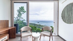 Una habitación moderna con un escritorio de madera, dos sillas, una mesita blanca y un ventanal con vistas al mar y la vegetación.