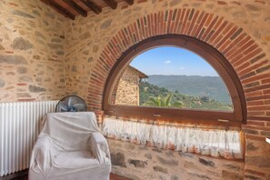Una habitación con paredes de piedra, un amplio ventanal arqueado, un radiador y una silla cubierta.
