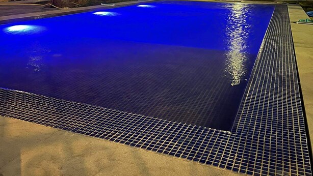A swimming pool with a blue illuminated edge, surrounded by palm trees and a building in the background.
