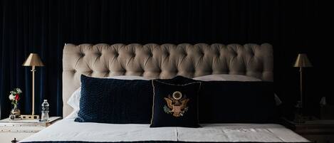 A bedroom with a tufted headboard, a dark blue quilted bedspread, and two pillows, one with an embroidered emblem.