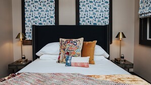 Un lit bien fait, avec du linge de lit blanc, un coussin coloré et un plaid à motifs. Il y a deux tables de chevet, chacune avec une lampe et une bouteille d’eau. La pièce est dotée d’une tête de lit sombre et d’œuvres d’art encadrées au-dessus du lit.