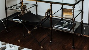 Una habitación con una mesa de vidrio, dos sillas negras, un adorno metálico en la pared y dos fotografías enmarcadas.