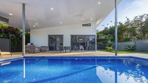 Un área de piscina moderna al aire libre con una piscina azul cristalina, una zona de estar cubierta con sillas y una mesa, y vistas de la vegetación circundante.
