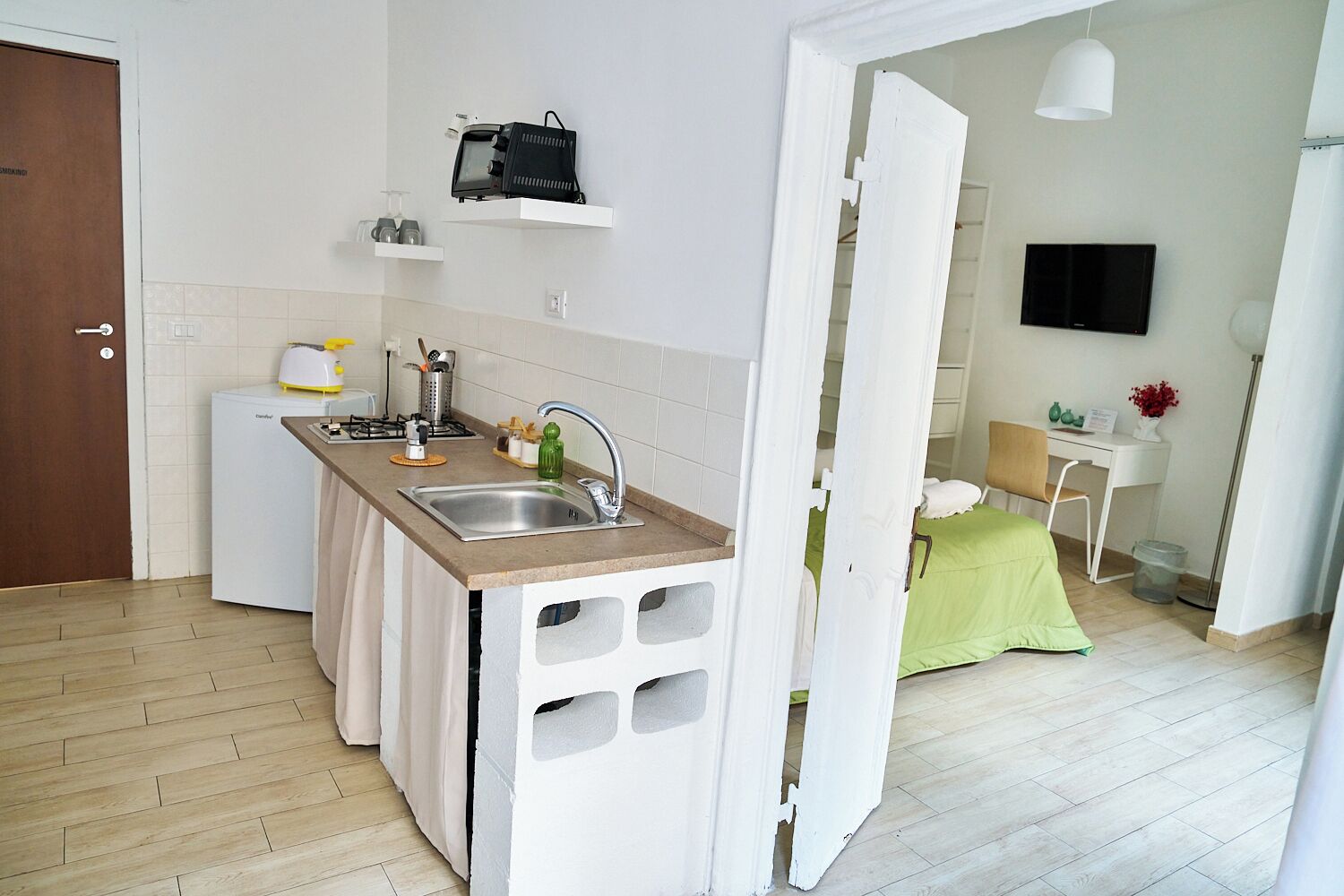 A compact kitchen with a sink, countertop, and a small refrigerator. There is a white door on the left, a white wall with a mounted television, and a green bed in the background.