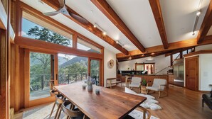 Un vaste espace à manger avec une table et des chaises en bois, de grandes fenêtres offrant une vue sur les montagnes et une cuisine à l’arrière-plan.