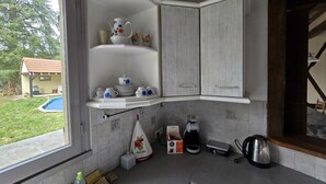 A kitchen with white cabinetry, a built-in shelf with white and blue floral dishware, a coffee maker, a kettle, and a small window overlooking a backyard. - Gîte du Puits Tournant (Poilly-lez-Gien)