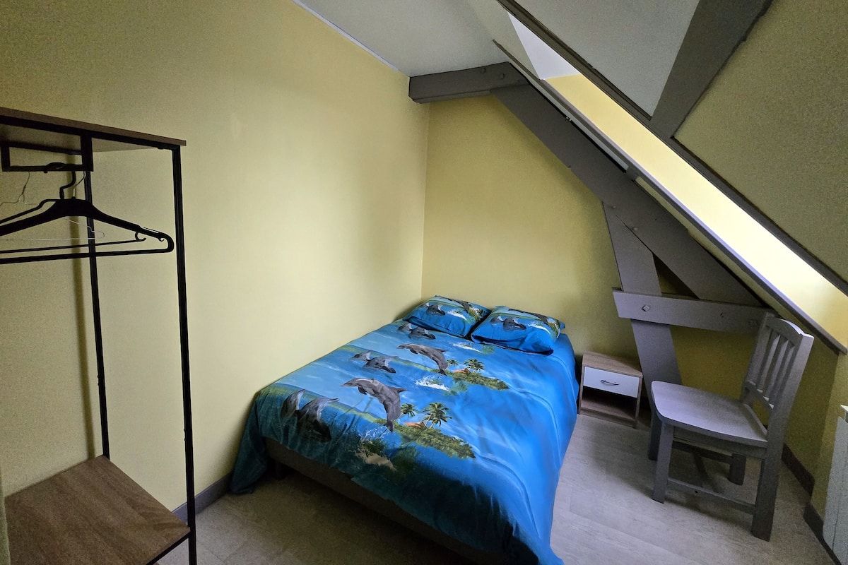 Une petite chambre avec un lit simple, un plafond en pente, une chaise en bois et un portant à vêtements.