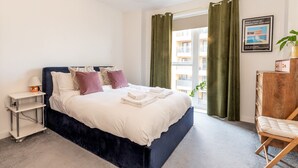A modern bedroom with a large bed, a bedside table, a lamp, a chair, and a view of the outside. - Stylish Flat Near Charlton Station - Pass the Keys (London)