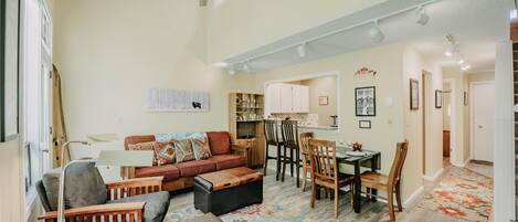 A spacious living area with a sofa, ottoman, and dining table. There is a ceiling fan, recessed lighting, and a patterned rug on the floor.