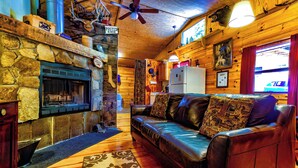 L’intérieur chaleureux d’un chalet avec une cheminée en pierre, un canapé en cuir, des murs en bois et un ventilateur de plafond.