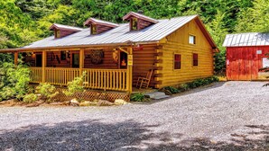 Une cabane en rondins avec une véranda, un abri de jardin rouge et un paysage boisé en arrière-plan.