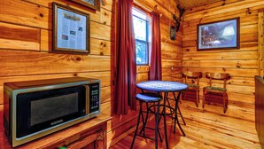 L’intérieur d’une cabane en bois est aménagé avec un four à micro-ondes, une petite table avec deux chaises, une affiche informative encadrée et une fenêtre avec des rideaux rouges.