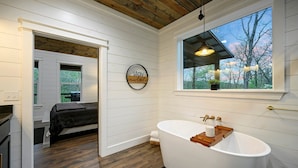 A modern bathroom with a freestanding bathtub, a large window with a view of trees, and a circular wall decoration with the text "stay awhile".