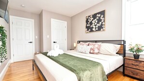 Un dormitorio con cama, dos puertas blancas, una obra de arte colgada en la pared, una mesita de noche con una planta y suelo de madera.