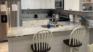 Una cocina moderna con armarios blancos, un refrigerador de acero inoxidable, un microondas y un lavamanos. Hay dos sillas blancas con asientos negros y un adorno de pared con tortugas marinas.