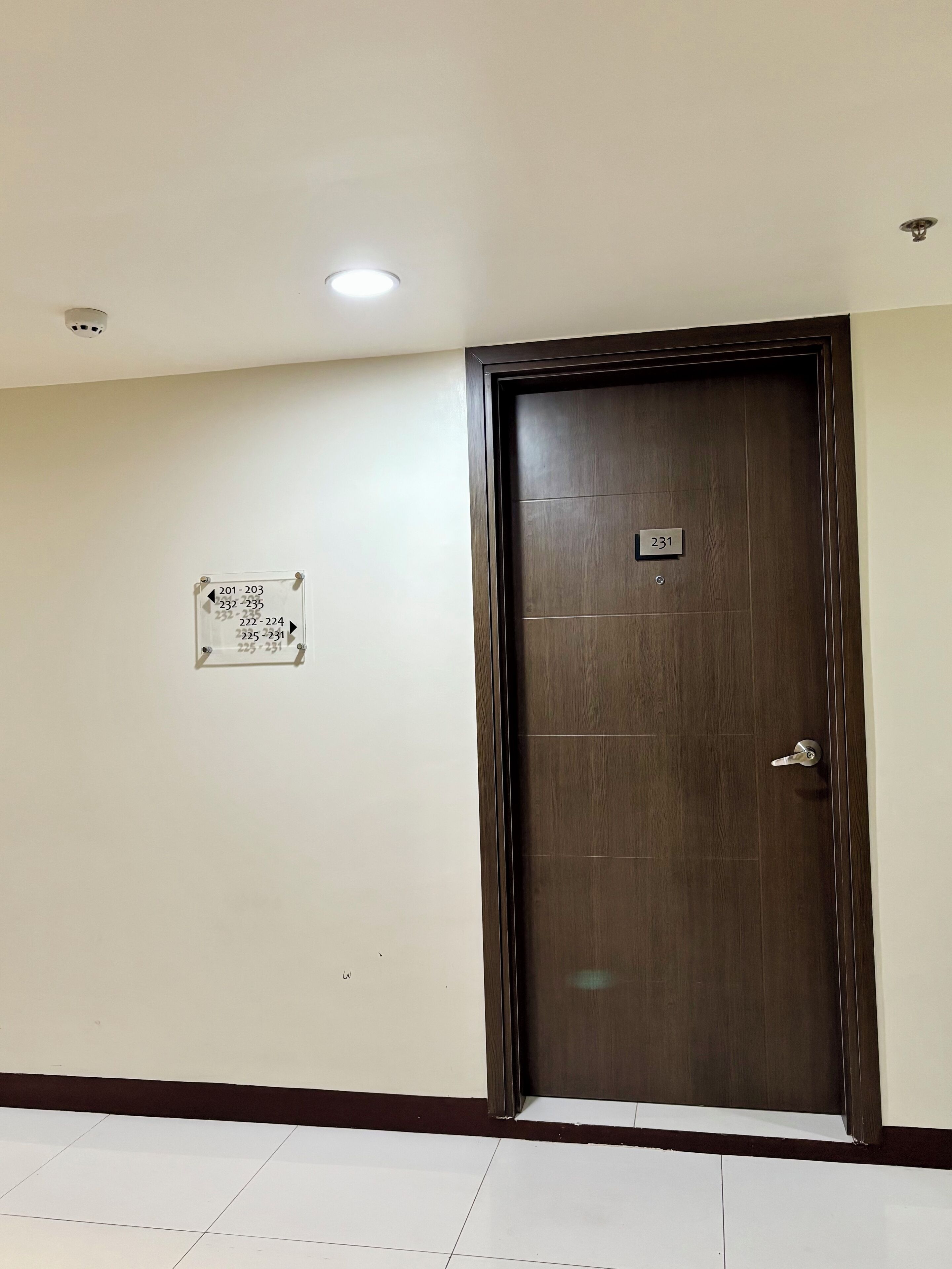 A closed wooden door with a number plate, a wall-mounted sign with room numbers, and a tiled floor.