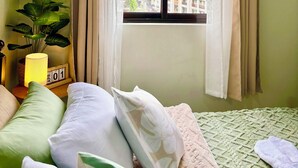 A bedroom with a bed, pillows, a bedside lamp, a potted plant, and a window with curtains.