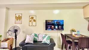 A living room with a black leather sofa, a fan, a TV mounted on the wall, and a dining area with a table and chairs.