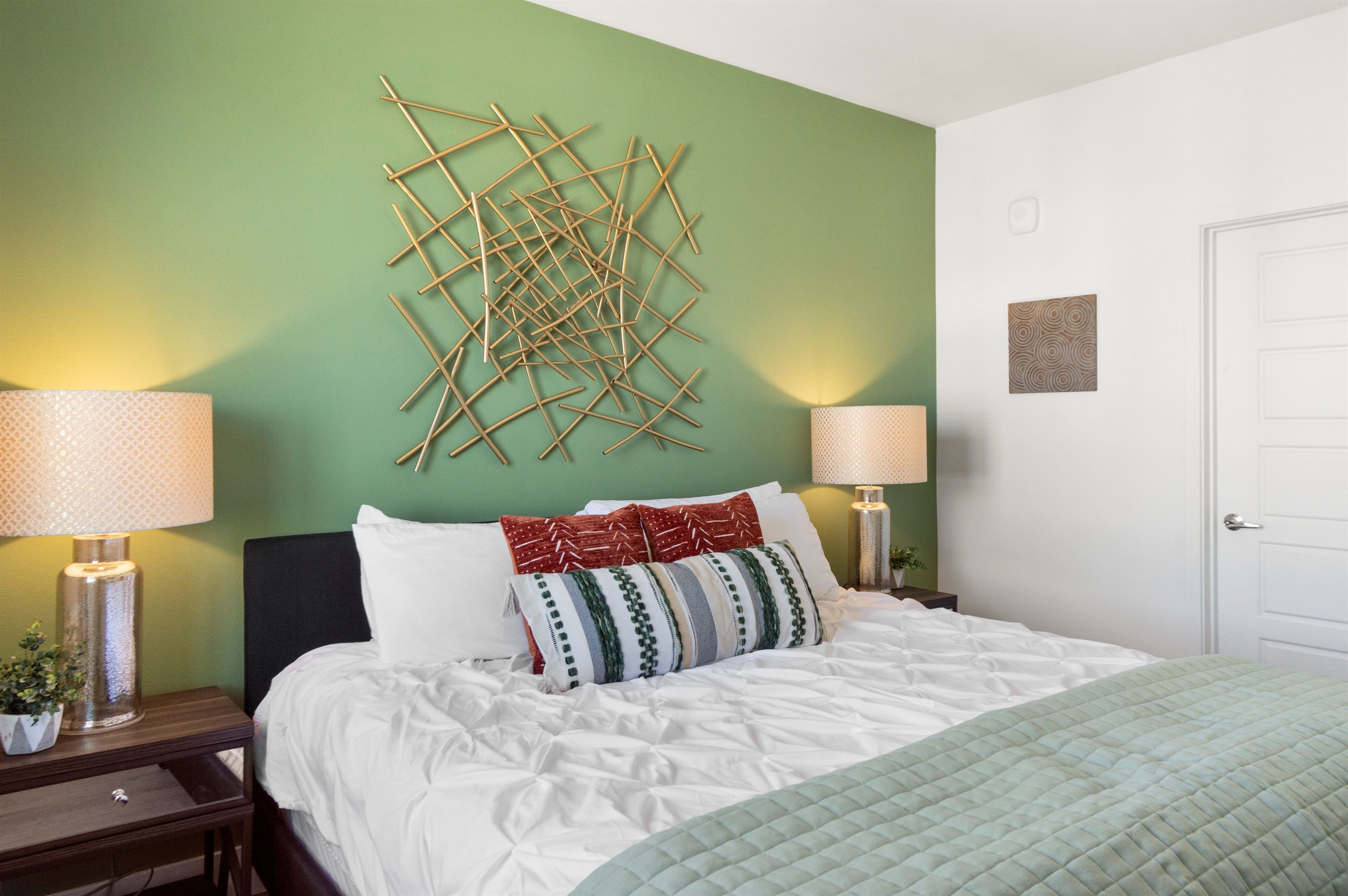 A neatly made bed with white bedding and a green quilt, two bedside lamps, a wall art piece, and a door.