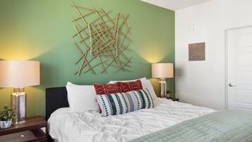 A neatly made bed with white bedding and a green quilt, two bedside lamps, a wall art piece, and a door.