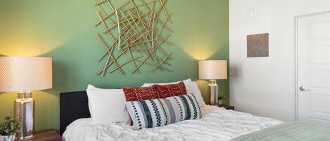 A neatly made bed with white bedding and a green quilt, two bedside lamps, a wall art piece, and a door.