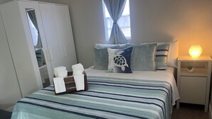 A neatly made bed with striped bedding, a bedside lamp, and a nightstand with a decorative item. A wardrobe is visible to the left, and a window with curtains is above the bed.