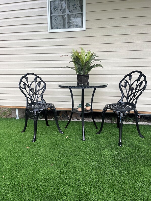 A small outdoor seating area with two black metal chairs and a round table, surrounded by artificial green grass.