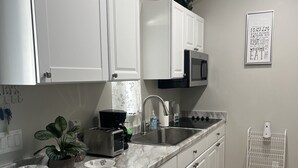 A modern kitchen with white cabinets, a marble countertop, a sink, and a microwave. There are stools and a plant on the counter.