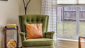 A green armchair with a cushion, a small wooden side table with books, a floor lamp, and a window with a view of a wooden fence. - Charming house minutes from everything in Monroe/West Monroe! 5-Star hosts! (West Monroe)