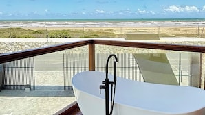 Un baño moderno con una bañera independiente sobre un piso de madera, con vistas a una playa y un cielo despejado.