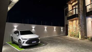 Um carro branco estacionado em área externa bem iluminada, com piso pavimentado, parede de pedra e um edifício com fachada de pedra e detalhes em madeira.