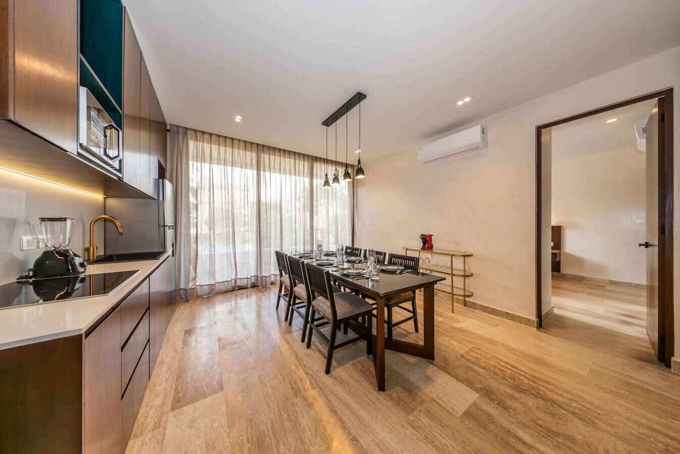 A modern kitchen with a built-in oven, a dining area with a wooden table and chairs, and a large window with sheer curtains.