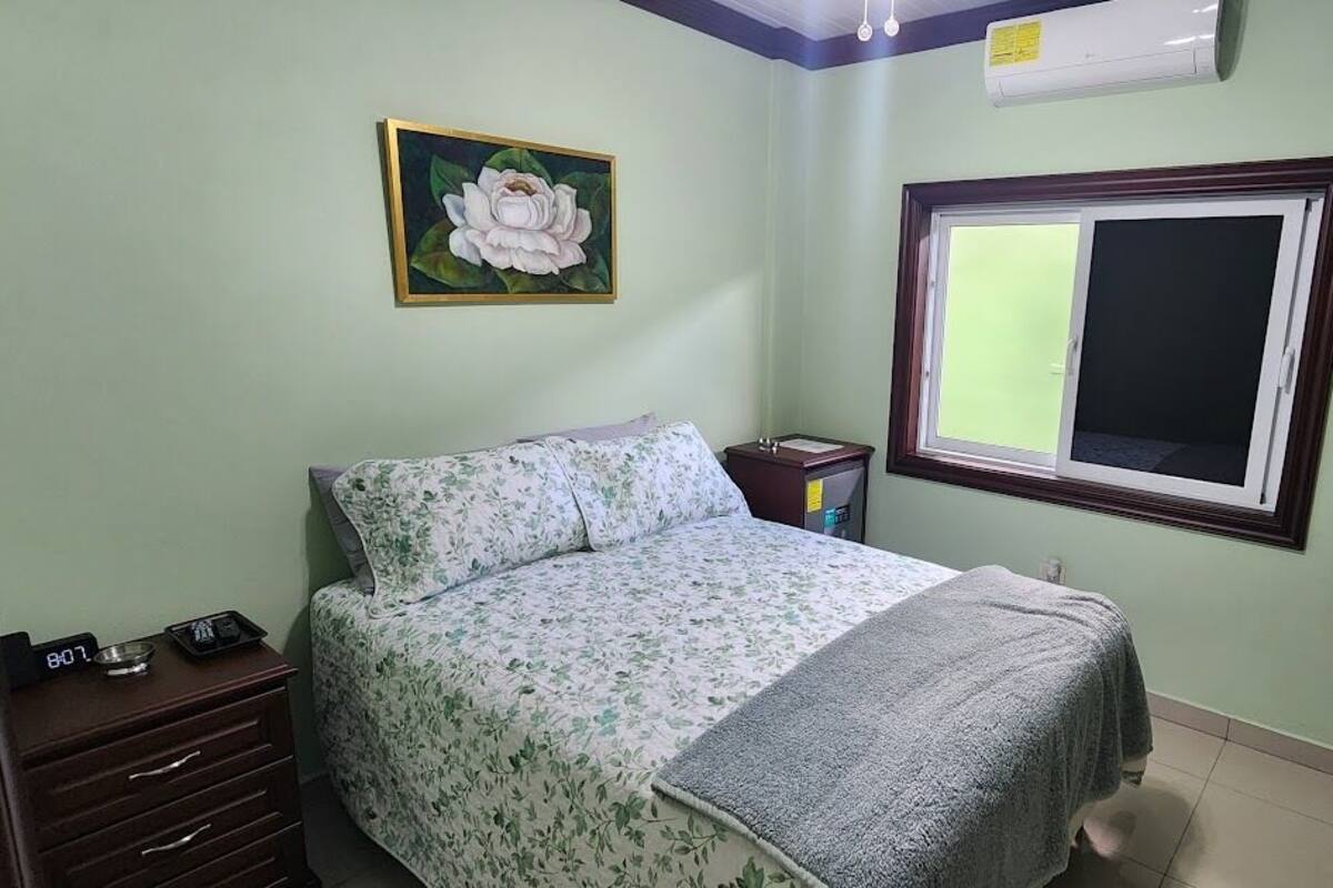 A bedroom with a bed, a nightstand, a wall clock, a window, and a painting.