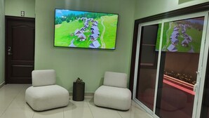 Un couloir avec deux fauteuils blancs, un téléviseur fixé au mur qui diffuse un paysage rural, et une porte avec une petite pancarte.