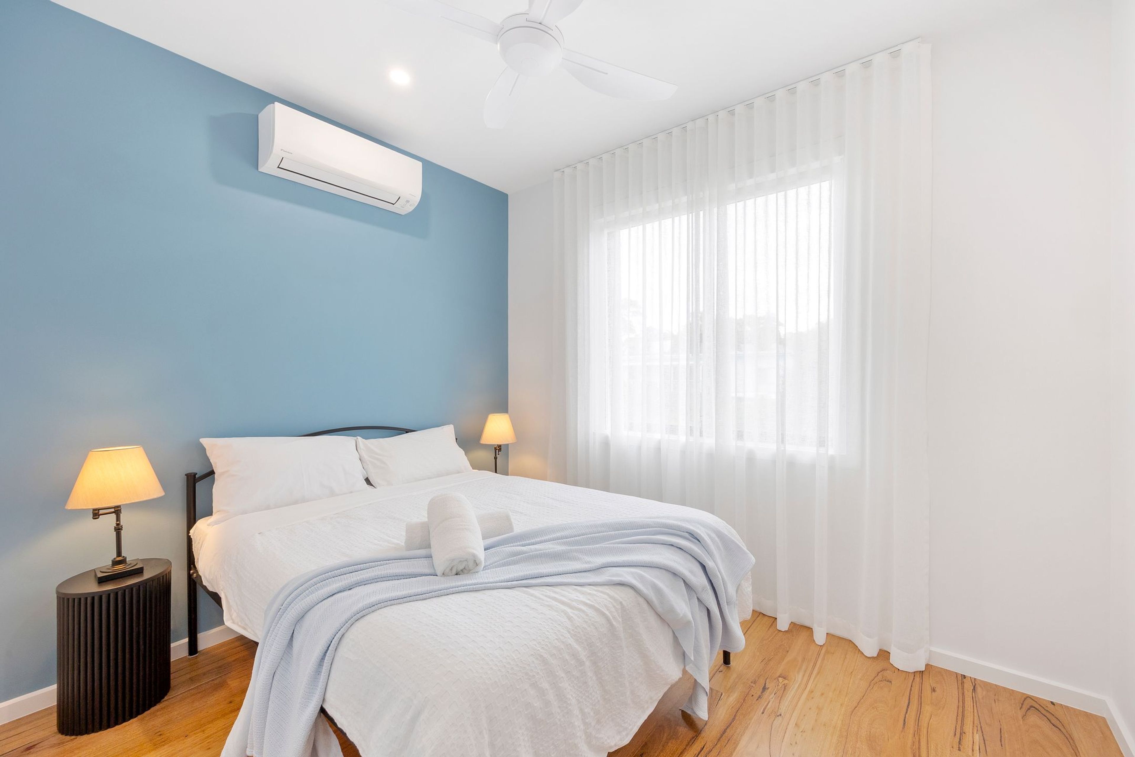 A hotel room with a bed, two bedside lamps, a ceiling fan, and a window with sheer curtains.