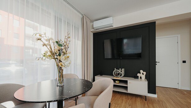 A modern living room with a round black table, beige chairs, a vase with white flowers, a flat-screen TV mounted on a black wall, and a white cabinet with decorative items.